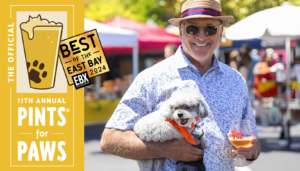 A smiling man in sunglasses and a hat holds a small white dog wearing a bandana and a Pints for Paws beer glass, standing in front of festival tents at the 11th Annual Pints for Paws event.